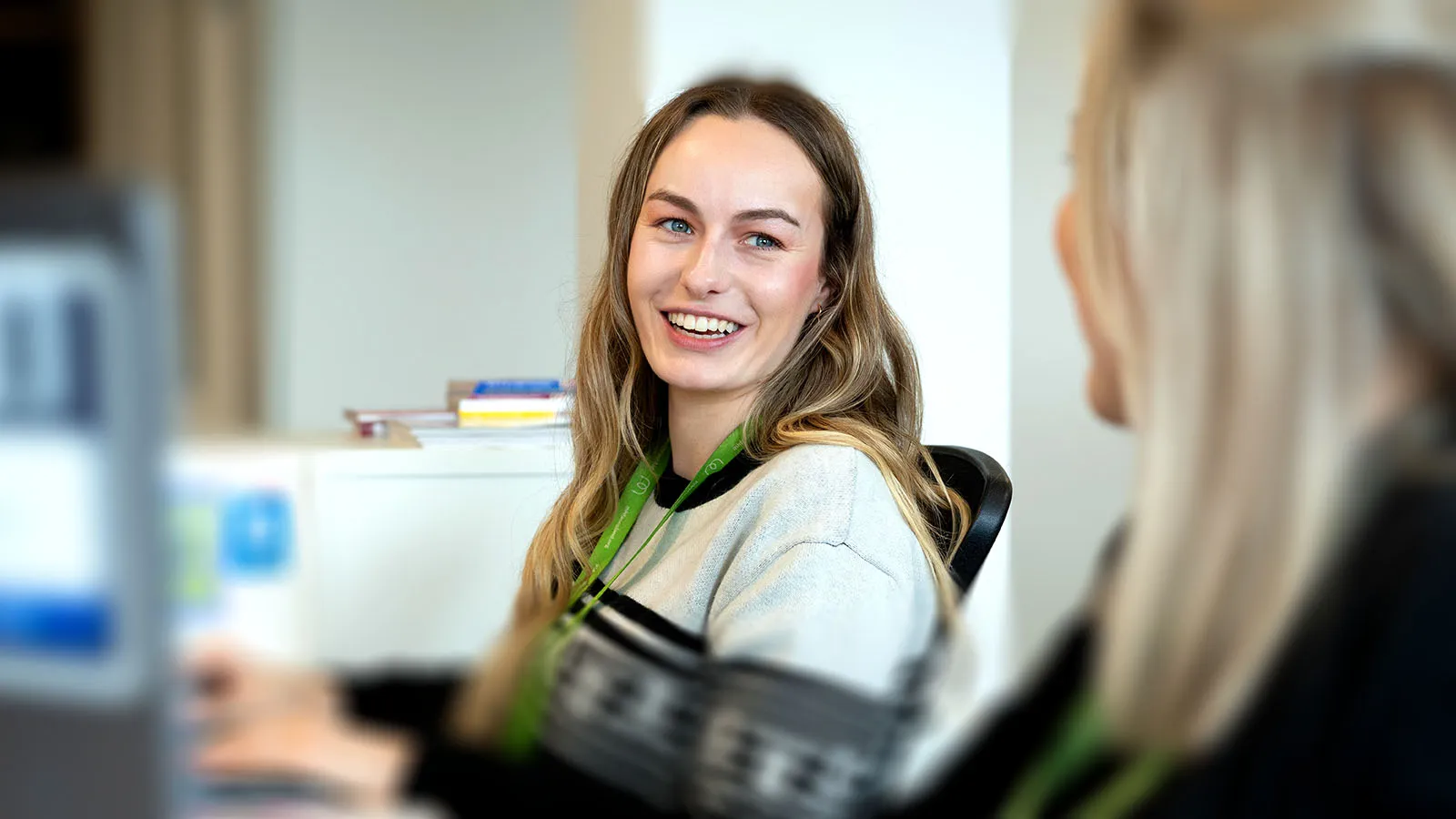 A woman with long hair, sitting at a desk, smiles towards the camera.
