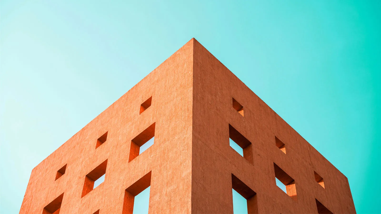 A minimalist orange building with square window openings, photographed from below against a bright blue sky.