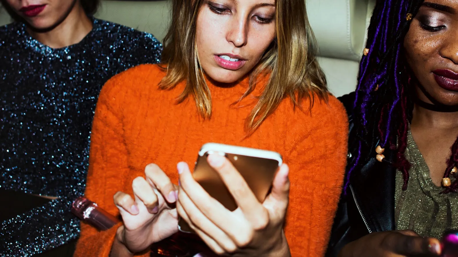A trendy woman with a blonde bob wearing an orange jumper, checks her phone in the back of a cab, a bottle of champagne resting on her lap.