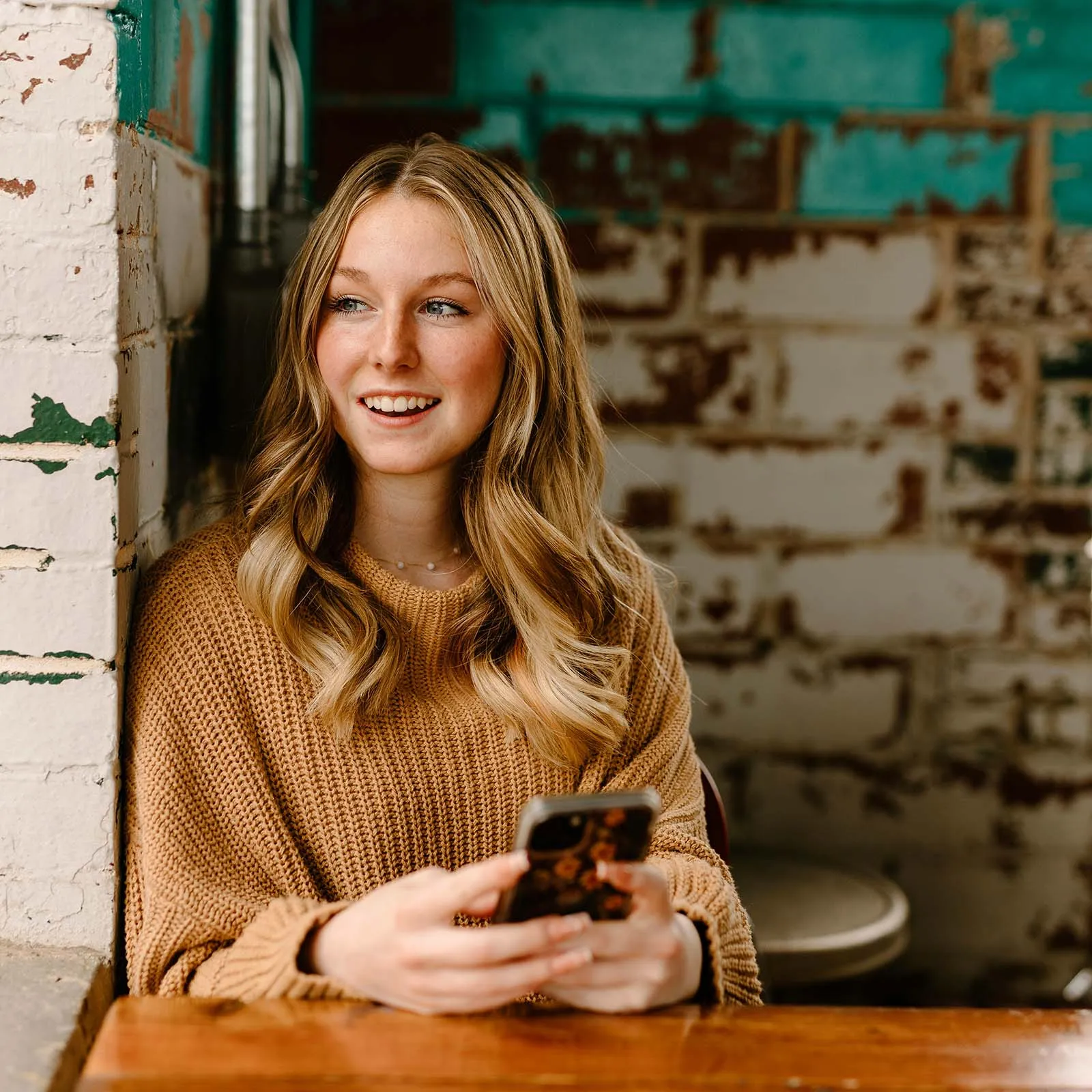 A young, blonde woman sits in a cafe, holding a phone and smiling.