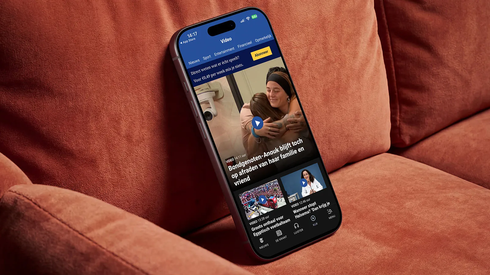A phone displaying the De Telegraaf news app rests on a brown sofa.
