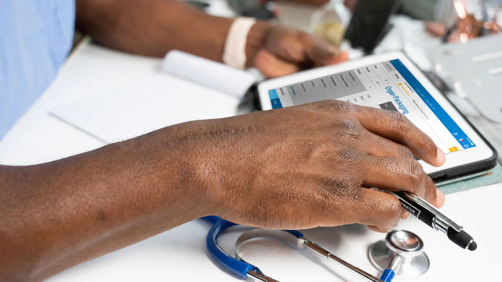 Close up of a stethoscope and hands resting on a table, touching a tablet displaying DonorPath NHS app.