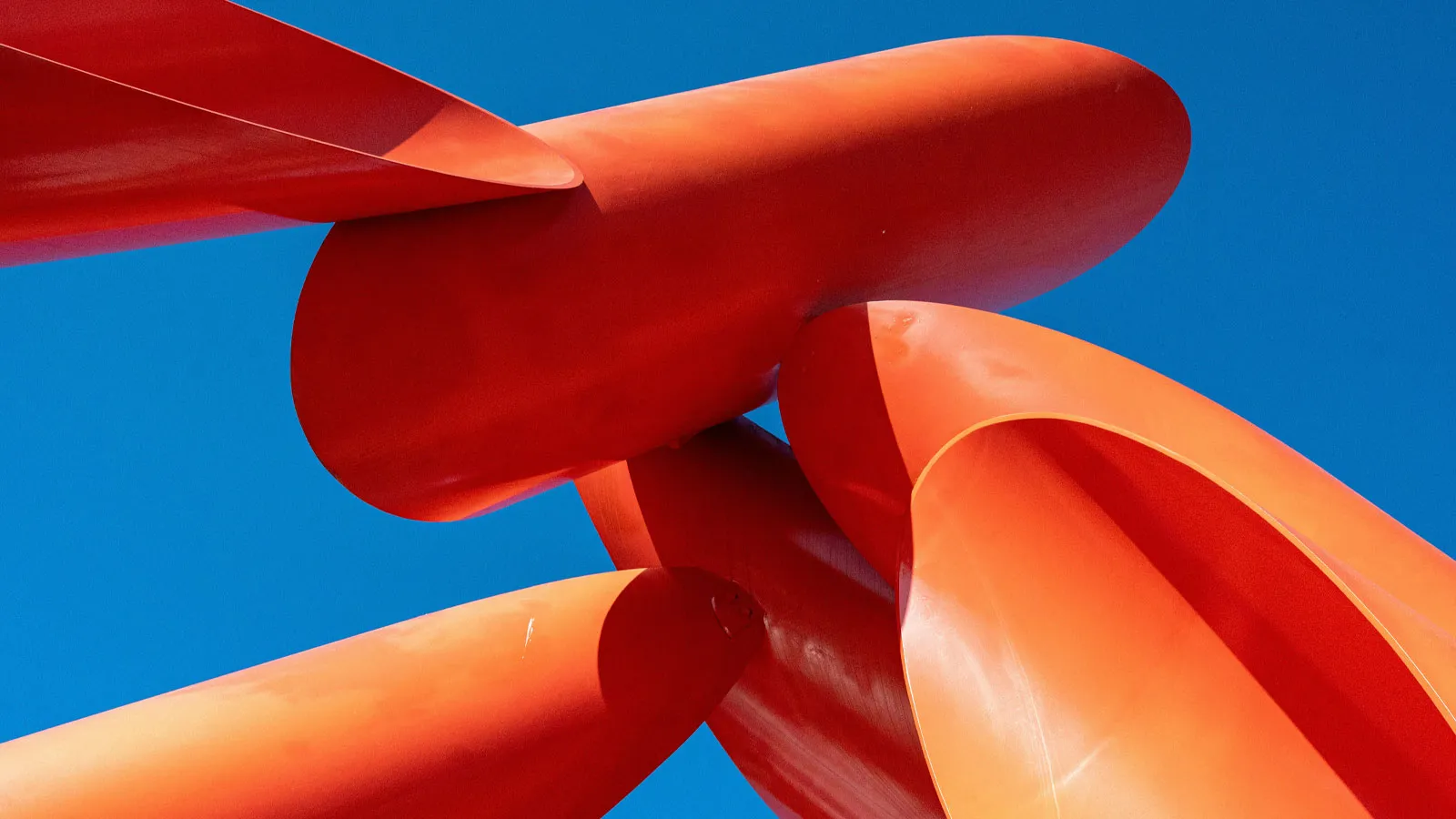 An abstract sculpture of smooth orange-red curved forms stands against a clear blue sky.