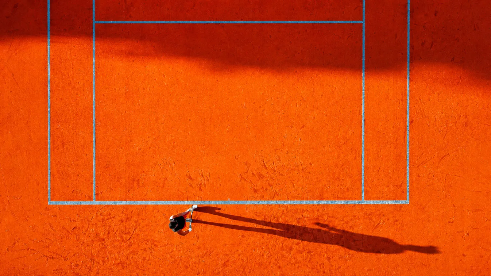 Aerial view of an orange tennis court, with a person about to serve, their shadow stretching across the clay.