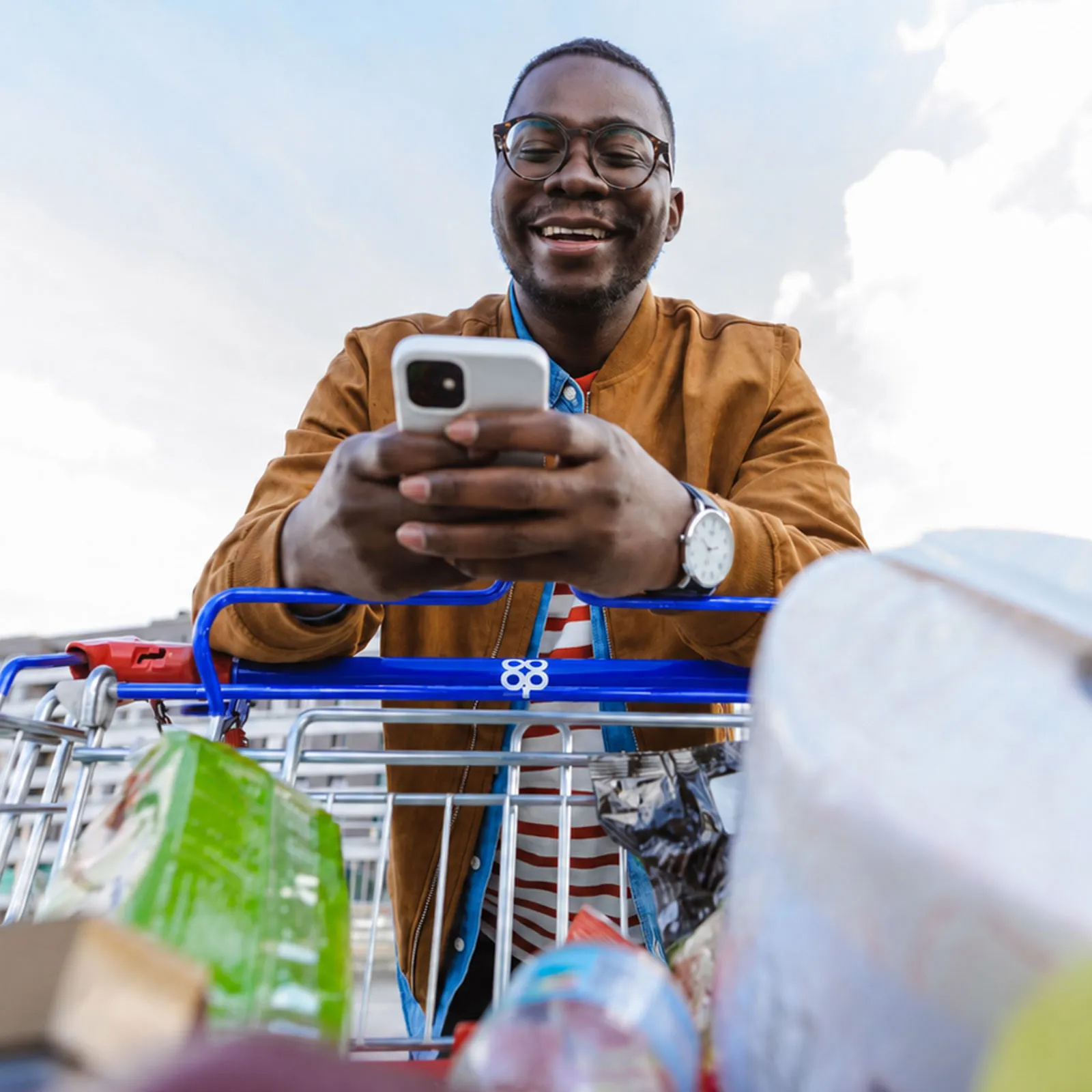 A man leans on the handle of a Co-op shopping trolley, smiling at his phone