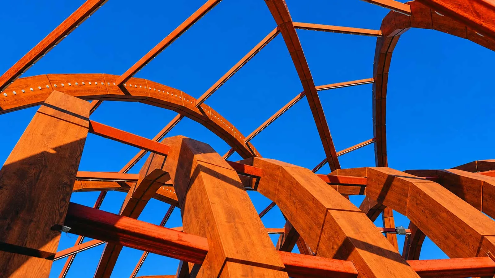 Upward view of an abstract steel framework with intersecting beams against a clear blue sky.