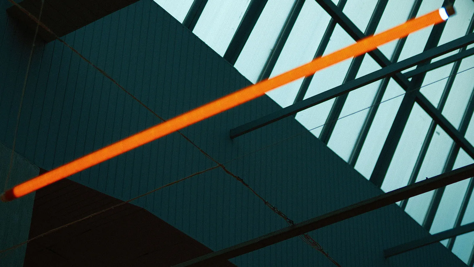 Orange illuminated light installation suspended diagonally inside a modern building with a glass roof and exposed steel beams.
