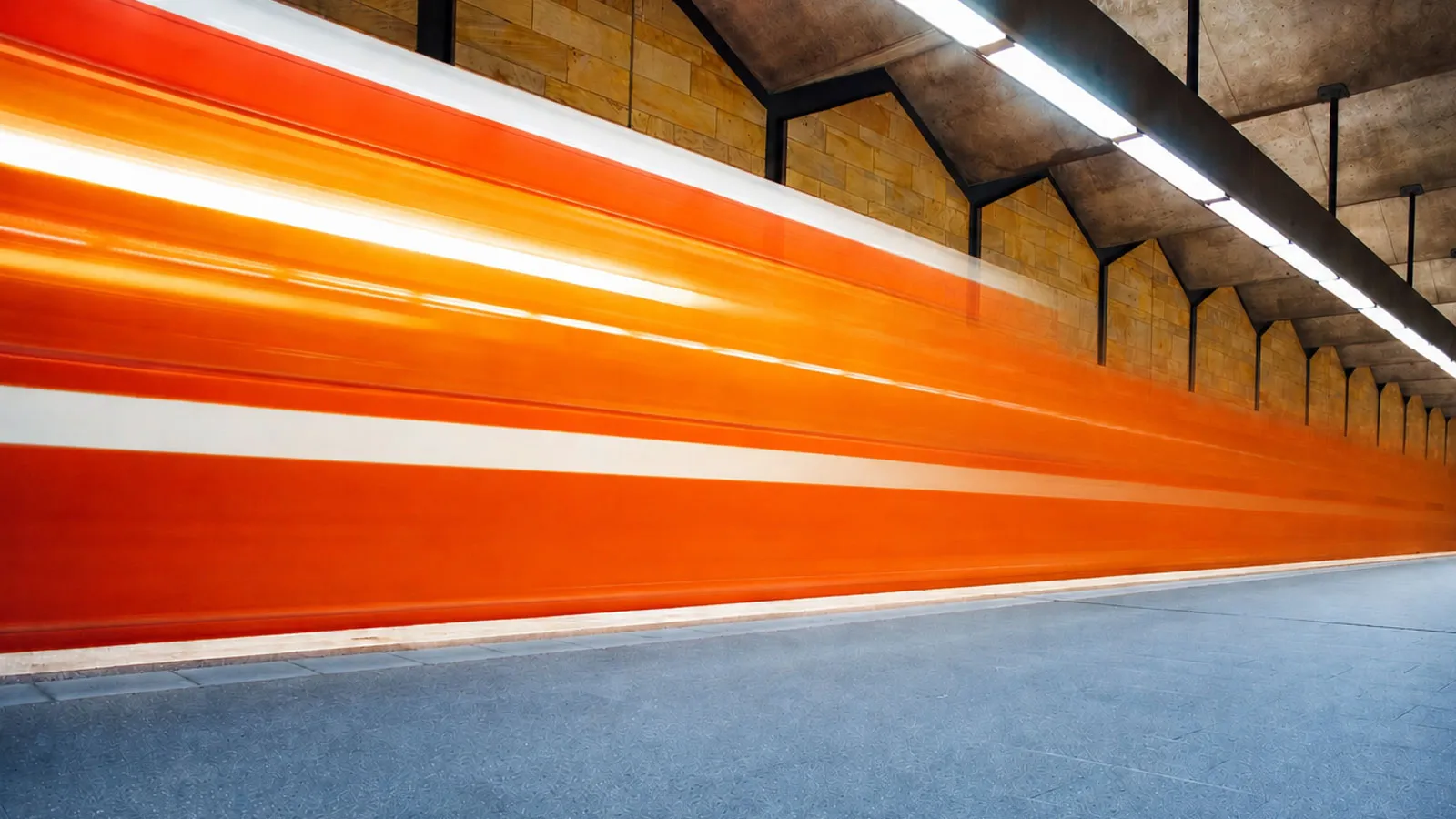 A blurred orange-and-white train speeds through a modern underground station with a blue-grey platform.