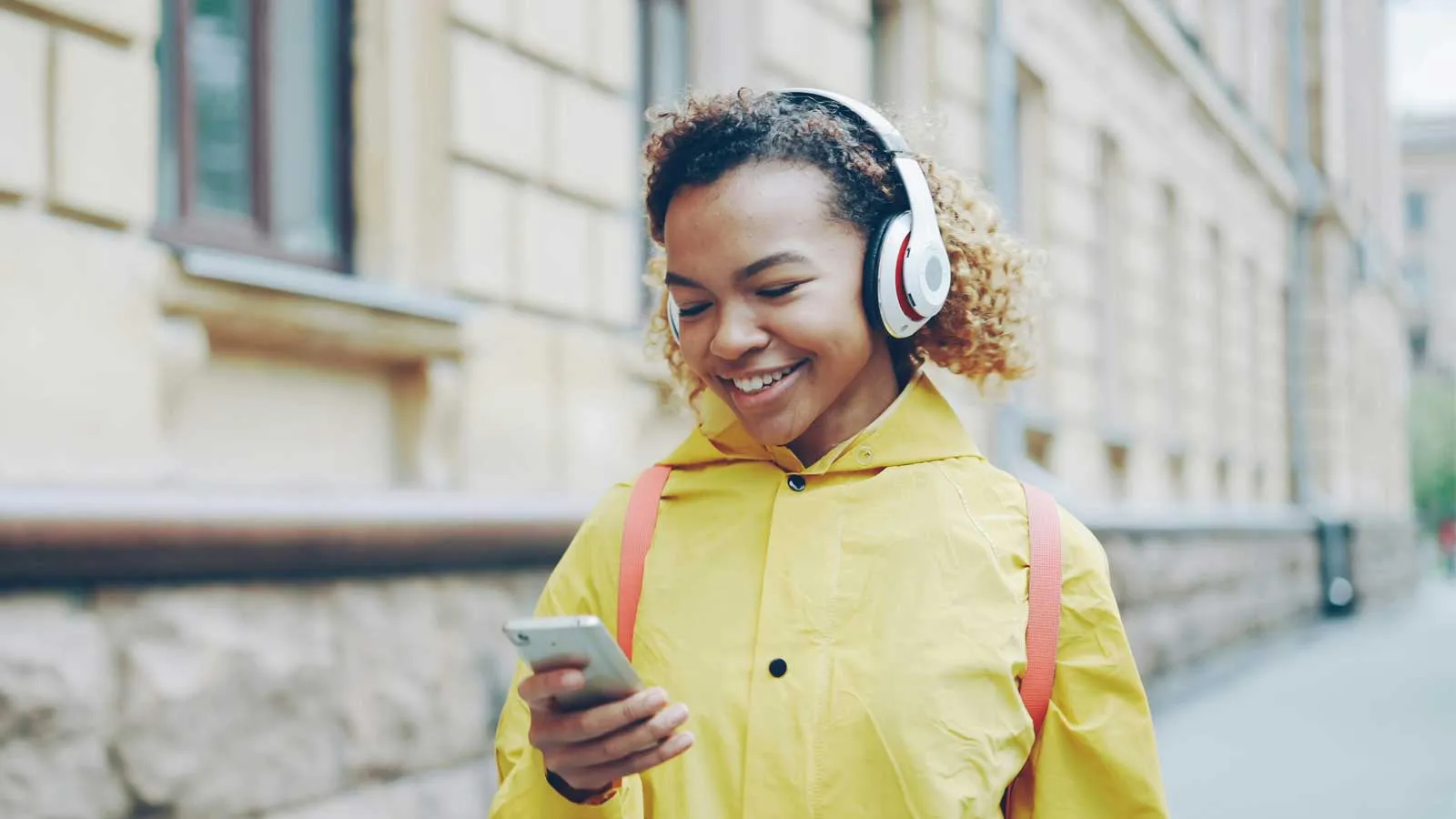 A girl wearing headphones and a yellow jacket smiles at her phone.