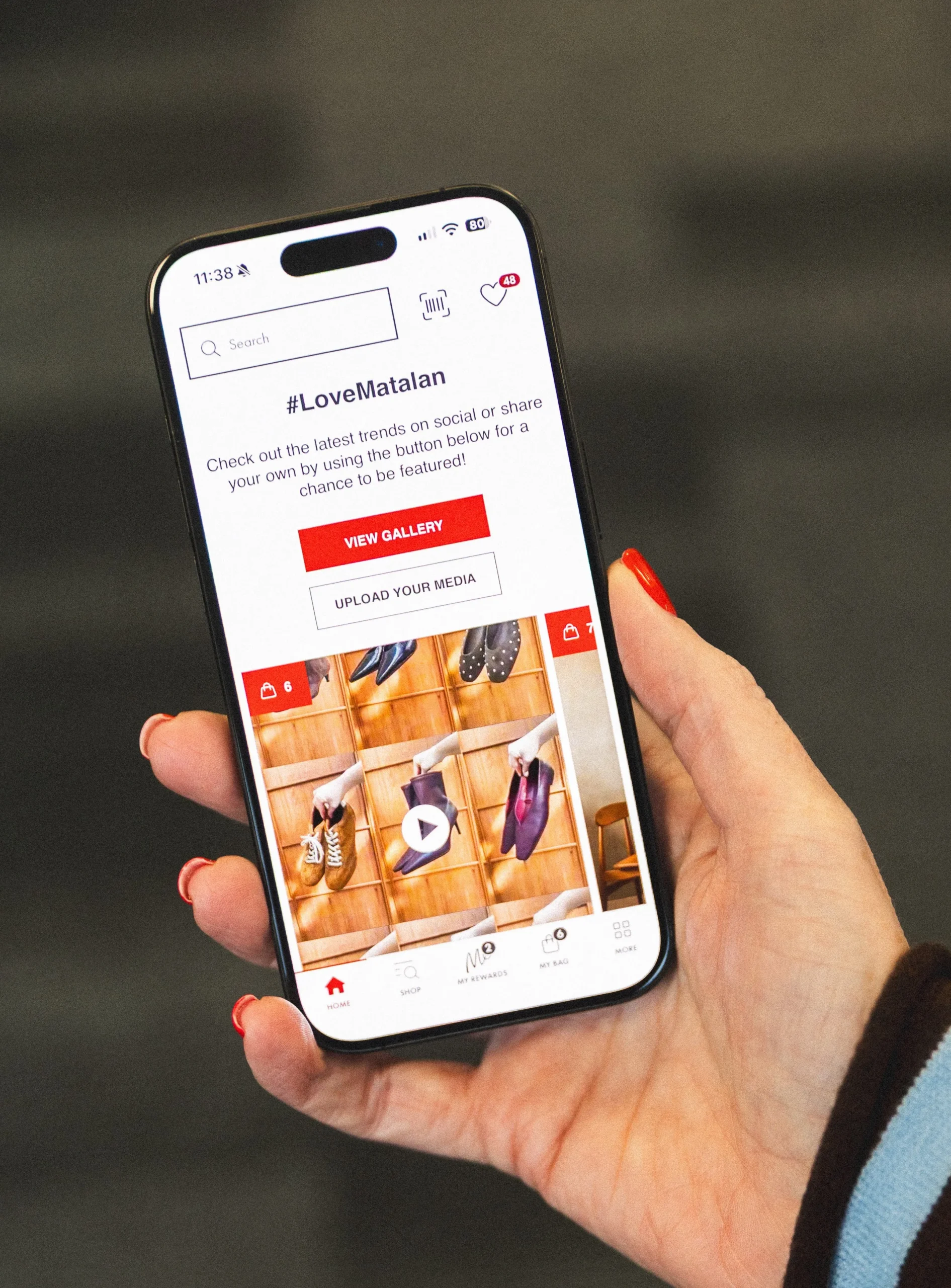 A hand with red nail polish holds a smartphone showing a fashion shopping app with a sale banner and clothing photos; striped sleeve against a dark background.