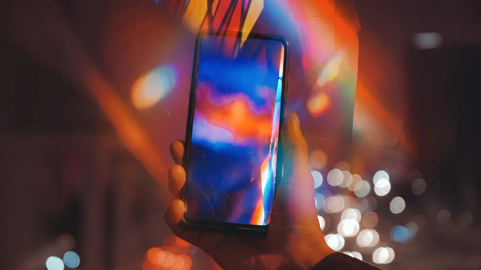 A hand holds a smartphone displaying vivid blue and orange colours against blurred city lights,