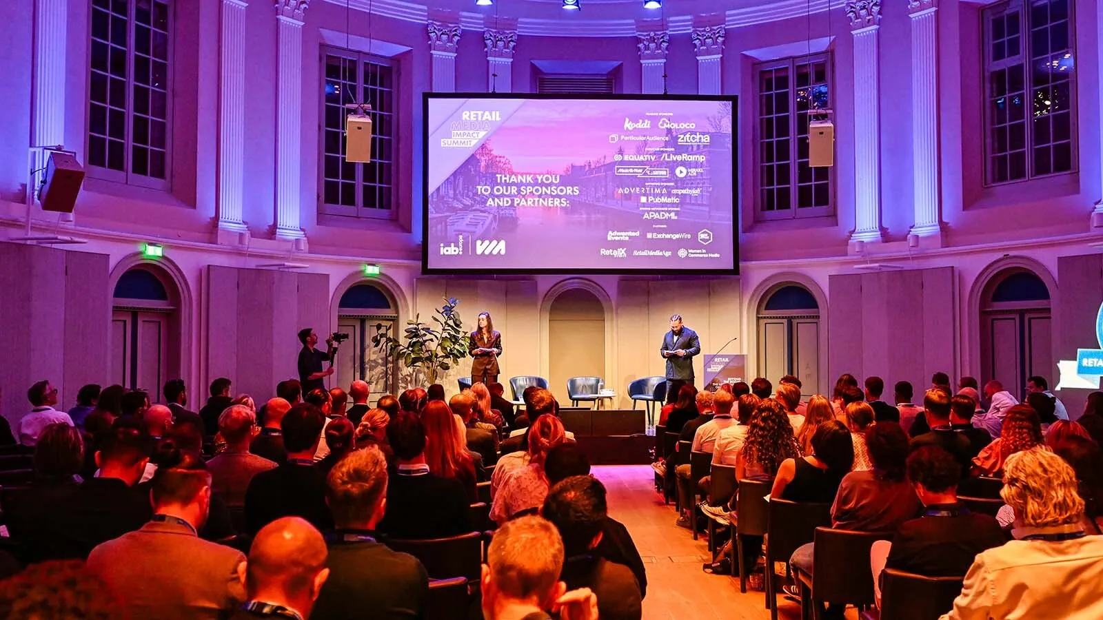 An audience watches a speaker beneath a sponsor thank-you screen in a grand, purple-lit hall,
